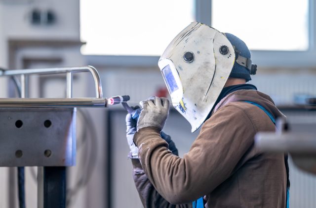 A male welder is using a welding machine to weld a metal piece in a building. He is wearing a welding helmet for personal protective equipment